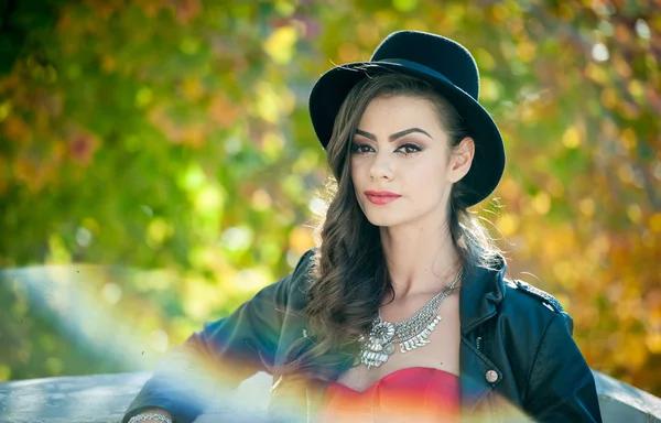 Beautiful woman with black hat posing in autumnal park. Young brunette spending time during autumn in forest. Long hair attractive girl with creative makeup and red dress, outdoors shot during fall