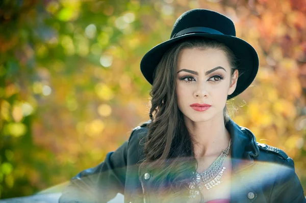 Beautiful woman with black hat posing in autumnal park. Young brunette spending time during autumn in forest. Portrait of long dark hair attractive girl with creative makeup, outdoors shot during fall