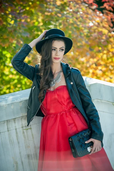 Beautiful woman with black hat posing in autumnal park. Young brunette spending time during autumn in forest. Long hair attractive girl with creative makeup and red dress, outdoors shot during fall