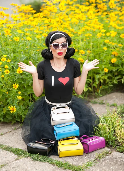 Beautiful girl with retro look wearing a black outfit having fun in park displaying a lot of colored bags. Fashionable brunette with sunglasses and purses in front, yellow flowers on background