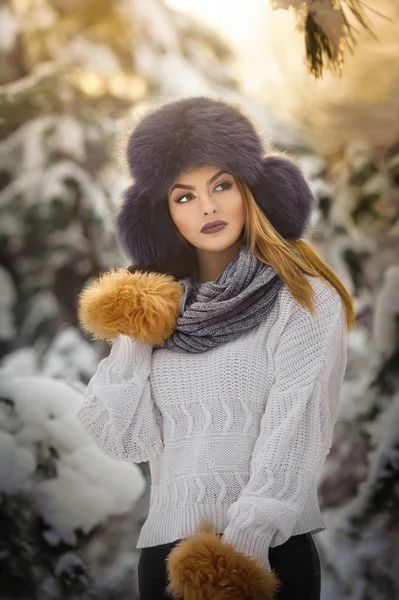 Beautiful woman in white pullover with over-sized fur cap enjoying the winter scenery in forest. Blonde girl posing under snow-covered trees branches.Attractive young female in bright cold day, makeup