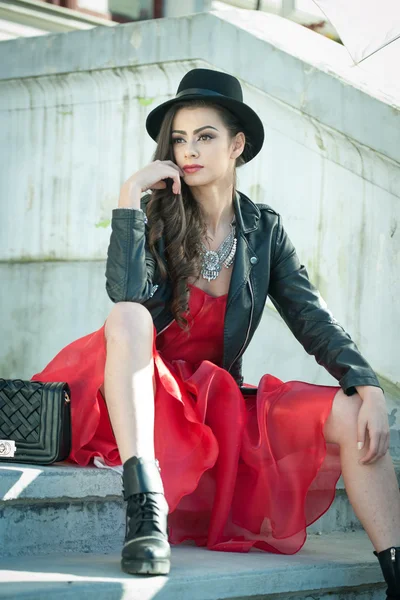 Beautiful woman with black hat, red dress and boots posing sitting on stairs. Young brunette spending time during autumn. Long hair attractive girl with creative makeup and red dress, outdoors shot