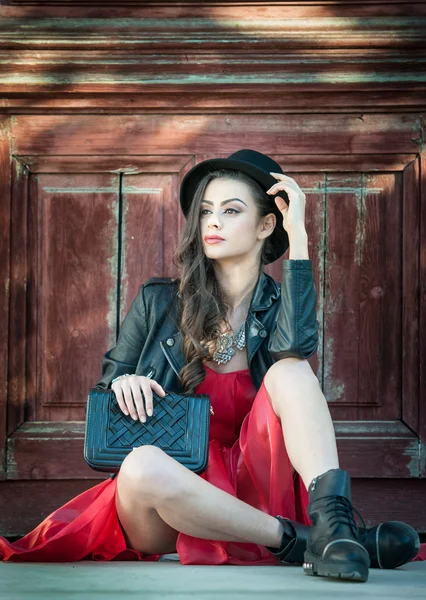 Young beautiful brunette woman with red short dress and black hat posing sensual in vintage scenery. Romantic mysterious young lady relaxing sitting on floor against vintage wooden wall