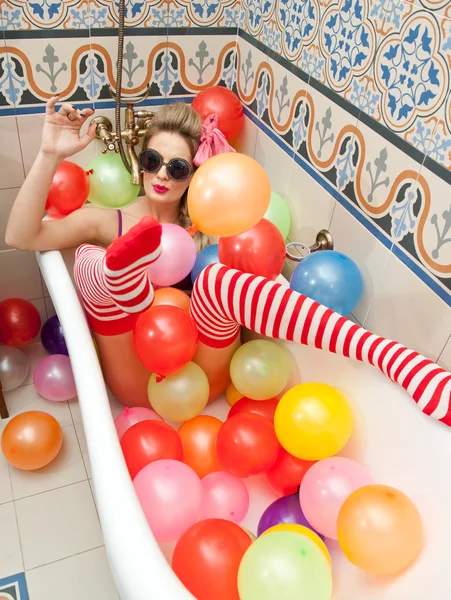 Blonde woman with sunglasses playing in her bath tube with bright colored balloons. Sensual girl with white and red striped stockings having fun in bathroom, covered with balloons
