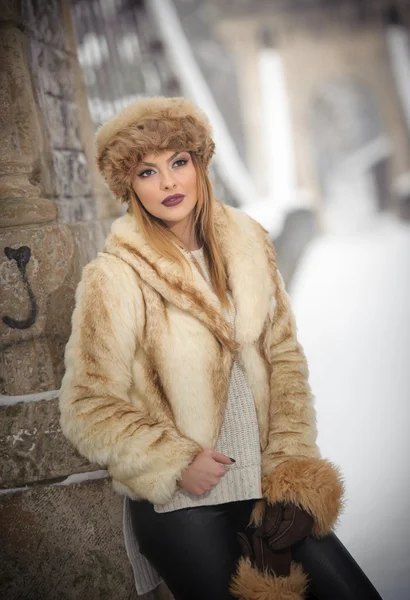 Attractive woman with brown fur cap and jacket enjoying the winter. Side view of fashionable blonde girl posing against snow covered bridge. Beautiful young female with cold weather outfit