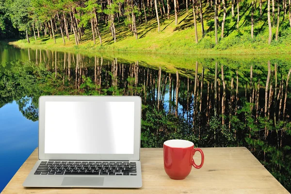 Laptop and red mug on wood table with forest pine