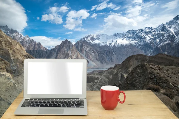Laptop and red mug on wood table with snow mountain in Pakistan