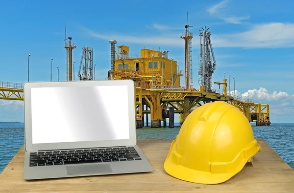 Laptop and yellow safety helmet on wood table with rig backgroun