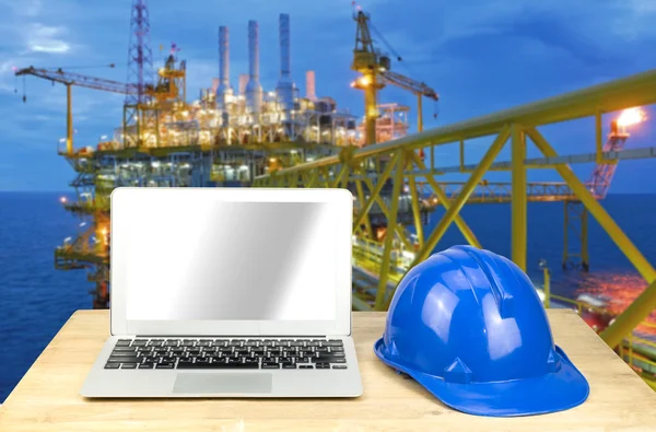 Laptop and blue safety helmet on wood table with rig background