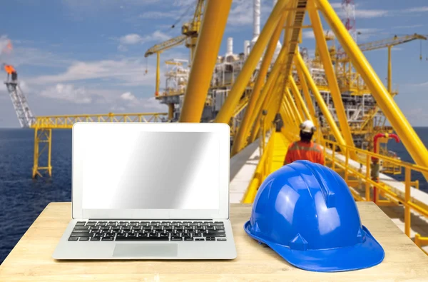 Laptop and blue safety helmet on wood table with rig background