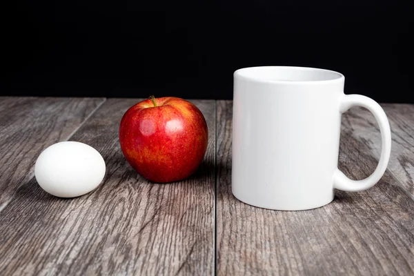 Hardboiled egg, red apple, and cup of coffee on a wood surface.