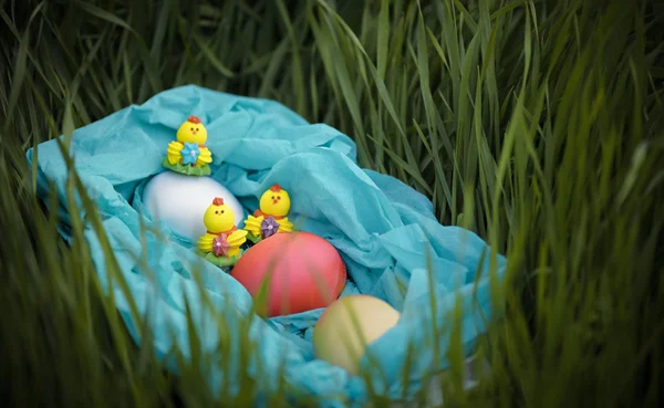 Three toy chickens and three easter eggs on the grass in a blue basket