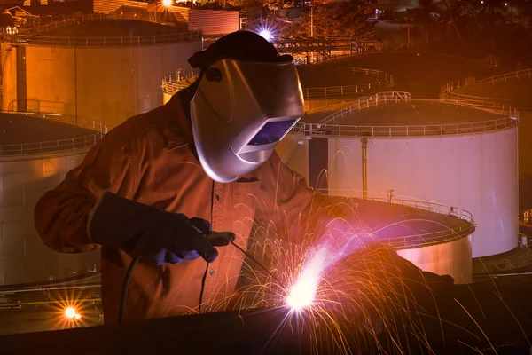 Industrial Worker with safety equipments and protective mask welding steel structure with Natural Oil and Gas storage tanks and in Petrochemical industrial plant at background