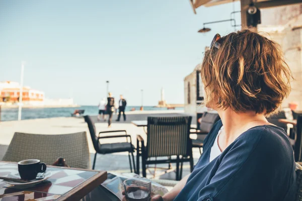 Young woman sitting in coffee bar and looking at old port in Chania, Crete, Greece