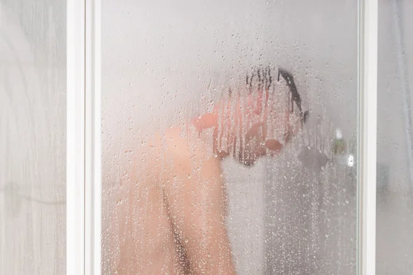 Male taking a shower standing behind transparent misted glass do