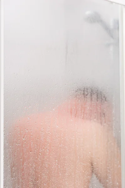 Portrait of young man taking a shower behind misted glass door