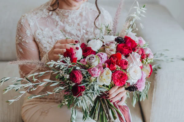 Wedding. Bouquet. Bride. Grain. Artwork. The bride in a white dress sitting on the couch and holding a bridal bouquet of red flowers, white flowers and greenery