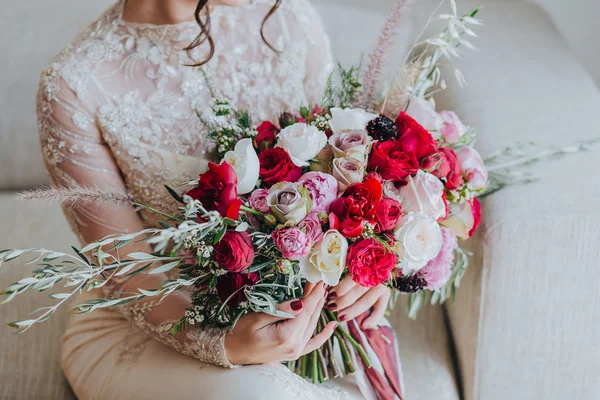 Wedding. Bouquet. Bride. Grain. Artwork. The bride in a white dress sitting on the couch and holding a bridal bouquet of red flowers, white flowers and greenery