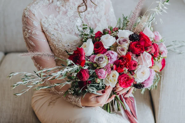 Wedding. Bouquet. Bride. Grain. Artwork. The bride in a white dress sitting on the couch and holding a bridal bouquet of red flowers, white flowers and greenery