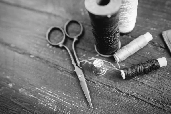 Sewing accessories: bobbins of thread, scissors, needle, thimble on wooden table. Black and white photo. Tailoring and sewing concept.