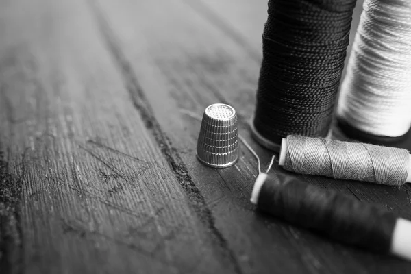Sewing accessories: bobbins of thread, needle, thimble on wooden table. Black and white photo. Tailoring and sewing concept.