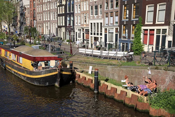 People relax on canal embankment near houseboat in Amsterdam