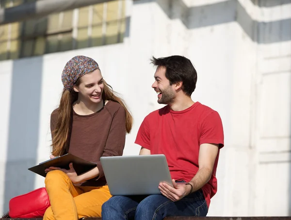 Two college students talking and working outdoors on laptop