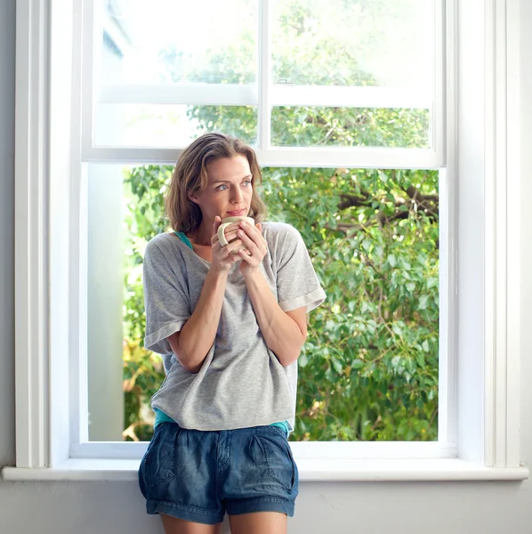 Healthy woman standing by window at home with cup of coffee