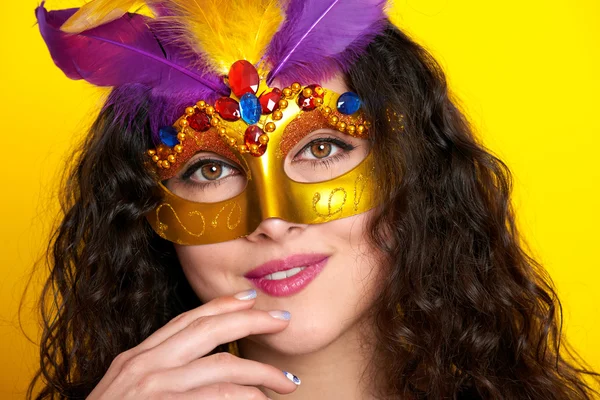 Woman face closeup in carnival masquerade mask with feather, beautiful girl portrait on yellow color background, long curly hair