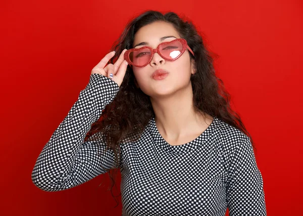 Young woman beautiful portrait flying kiss, posing on red background, long curly hair, sunglasses in heart shape, glamour concept