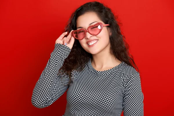 Woman winks, beautiful portrait, posing on red background, long curly hair, sunglasses in heart shape, glamour concept