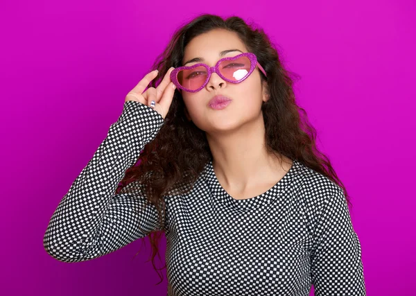 Young woman beautiful portrait flying kiss, posing on purple background, long curly hair, sunglasses in heart shape, glamour concept