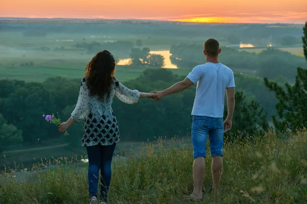 Romantic couple walk on hill at sunset on country outdoor, girl with bouquet of wild flower, beautiful landscape and bright yellow sky, love tenderness concept, young adult people