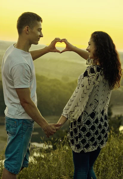 Romantic couple at sunset make a heart shape from hands, beautiful landscape and evening sky, love tenderness concept, young adult people, yellow toned