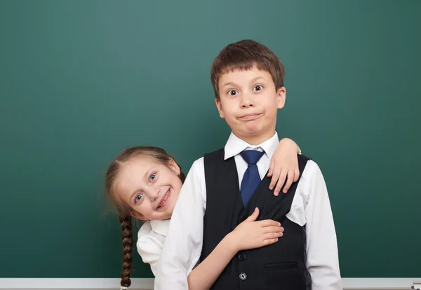 Two school student posing at the clean blackboard, grimacing and emotions, dressed in a black suit, education concept, studio photo