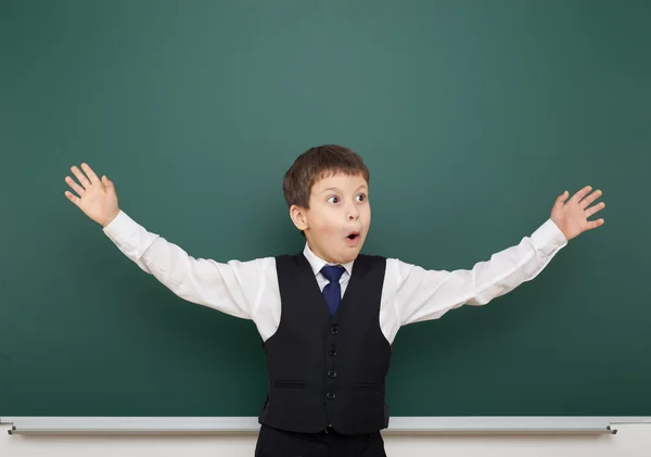 School student boy posing at the clean blackboard and open arms, grimacing and emotions, dressed in a black suit, education concept, studio photo