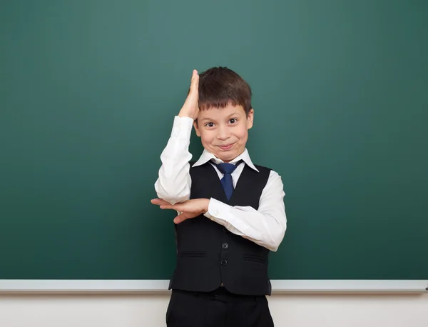 School student boy posing at the clean blackboard, grimacing and emotions, dressed in a black suit, education concept, studio photo