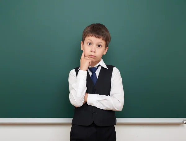 School student boy posing and think at the clean blackboard, grimacing and emotions, dressed in a black suit, education concept, studio photo