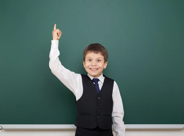 School student boy posing at the clean blackboard, show finger up and point, grimacing and emotions, dressed in a black suit, education concept, studio photo