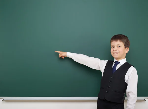 School student boy posing at the clean blackboard, show finger and point at, grimacing and emotions, dressed in a black suit, education concept, studio photo