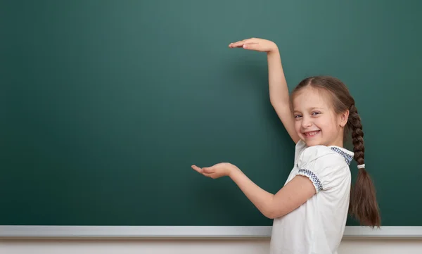 School student girl open arms at the clean blackboard, grimacing and emotions, dressed in a black suit, education concept, studio photo