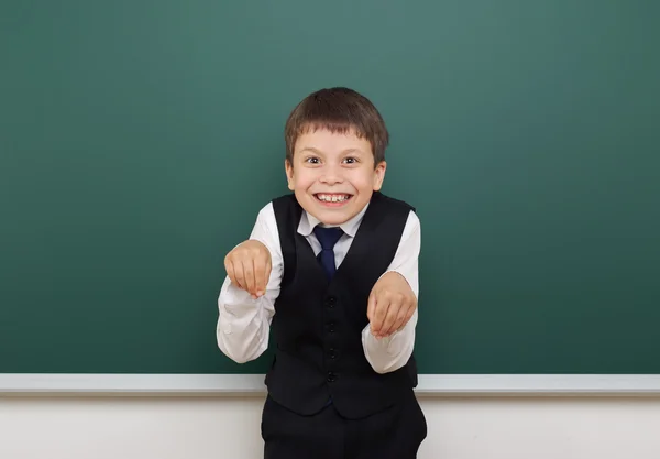School student boy posing at the clean blackboard, grimacing and emotions, dressed in a black suit, education concept, studio photo