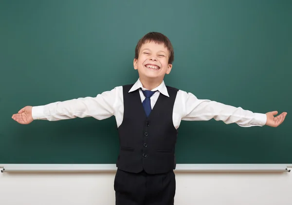 School student boy posing at the clean blackboard and open arms, grimacing and emotions, dressed in a black suit, education concept, studio photo