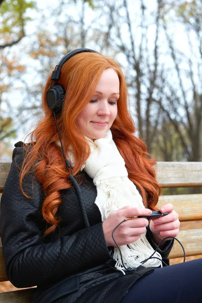 Girl listen music on audio player with headphones, sit on bench in city park, autumn season, yellow trees and fallen leaves