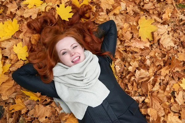 Redhead girl lying on leaves and wink in city park, fall season