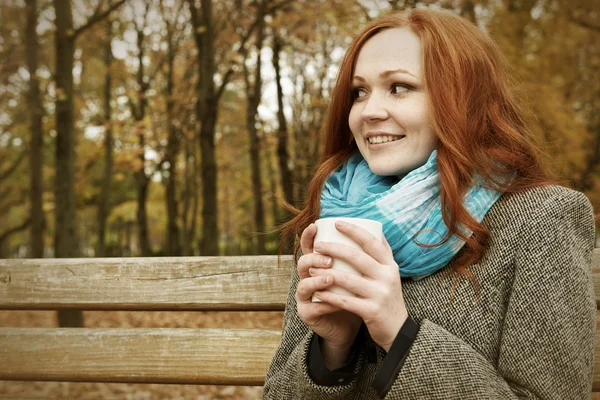 Happy girl with cup of tea sit on a bench in city park, fall season