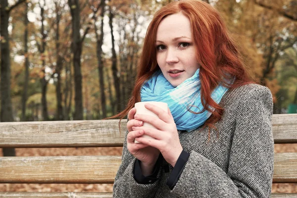 Redhead girl with cup of tea sit on a bench in city park, fall season