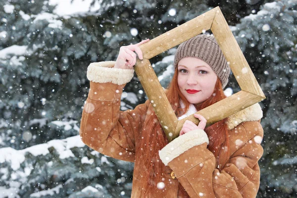 Woman outdoor portrait in wooden photo frame at winter season. Snowy weather in fir tree park.