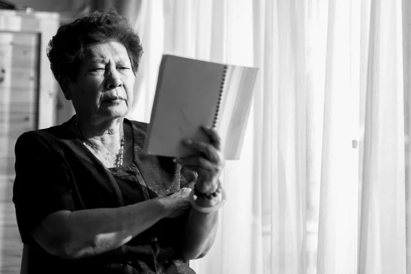 Black and white Asian senior woman reading a book with window li
