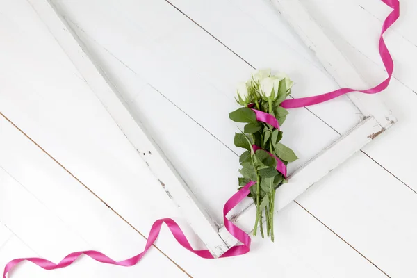 Five white roses with pink ribbon and old rustic window on white wooden floor